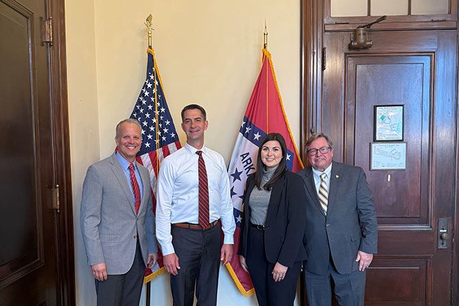 Arkansas Dealers Head to D.C.; Four people standing in front of U.S. and Arkansas flags in a formal setting. Three men and one woman are smiling, wearing suits, conveying professionalism.