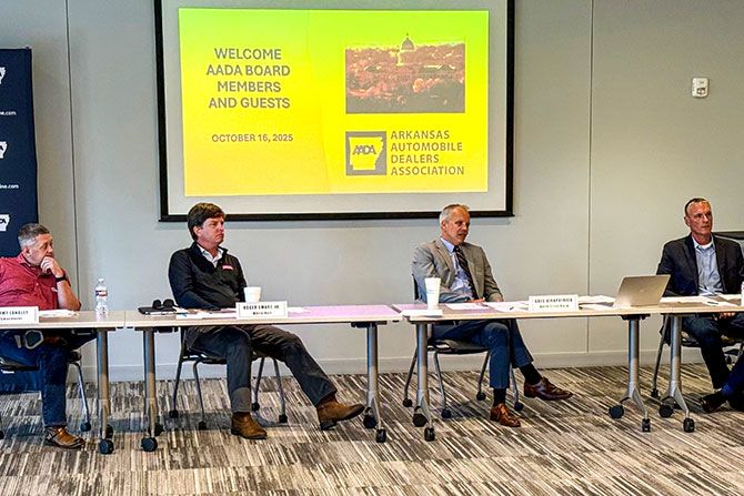 Fall Board of Directors Meeting Recap; A group of four men sitting at a conference table in a meeting room. A bright screen displays a welcome message for the Arkansas Automobile Dealers Association board meeting.