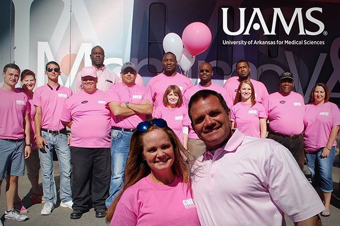 2026 TIME Dealer of the Year Nominee Jamie Cobb; A diverse group of people, mostly wearing pink shirts, smile and pose outdoors in front of a UAMS sign. The mood is cheerful and supportive.