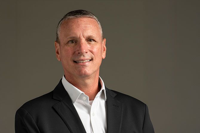 Getting to Know AADA Incoming Chair Michael Dickerson; Man in a black blazer and white shirt smiling warmly against a neutral gray background, conveying a professional and approachable demeanor.